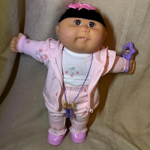 May include: A doll with black hair, a pink bow, and brown eyes wearing a white shirt with strawberry prints, a matching pink zip-up hoodie and trousers with strawberry patterns, a purple necklace with a small doll charm, and purple shoes.