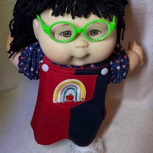Puede incluir: Una muñeca con cabello de lana negra, ojos marrones y gafas de plástico verde brillante. La muñeca viste una blusa azul marino con flores bajo un vestido de peto bicolor rojo y negro con un parche de arcoíris y manzana en el bolsillo.