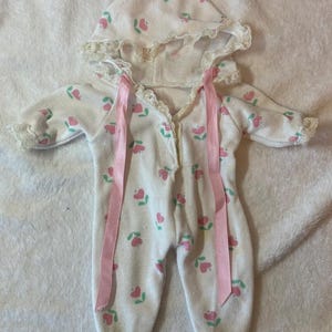 May include: A white hooded footed sleeper featuring a repeating pattern of small pink hearts with green stems. The garment includes long sleeves, a hood trimmed with white lace, and two long pink satin ribbons attached at the neckline.