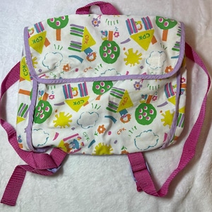 May include: A white backpack with a colorful pattern of trees, suns, and clouds. The backpack has pink straps and trim, with the letters "CPK" visible on the design. The flap is outlined in purple.