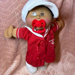 May include: A doll with a round face, one white eye, and one brown eye, wearing a white cap and a red sailor-style shirt with a white collar and an anchor emblem on the pocket. The doll has a bright red pacifier in its mouth and white shoes.