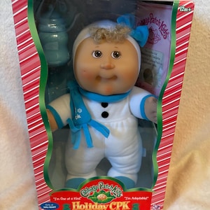 Pode incluir: Uma boneca Cabbage Patch Kids Holiday CPK com um traje de boneco de neve branco e azul, gorro e cachecol a condizer, embalada numa caixa às riscas vermelhas e brancas com o texto "I'm One of a Kind" e "I'm Adoptable" impresso na frente.