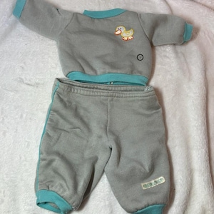 May include: A gray doll outfit featuring a sweatshirt and matching pants. The sweatshirt has a teal collar and cuffs, with a duck applique. The pants have teal side stripes and cuffs. The outfit is made of a soft fabric.