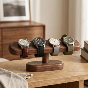Op de afbeelding: Een horlogestandaard van donkerbruin hout met een ovale voet en een horizontale balk die vier polshorloges vasthoudt. De horloges hebben diverse bandjes, waaronder bruin leer, zwart rubber, zilverkleurig mesh en een olijfgroene stoffen band.