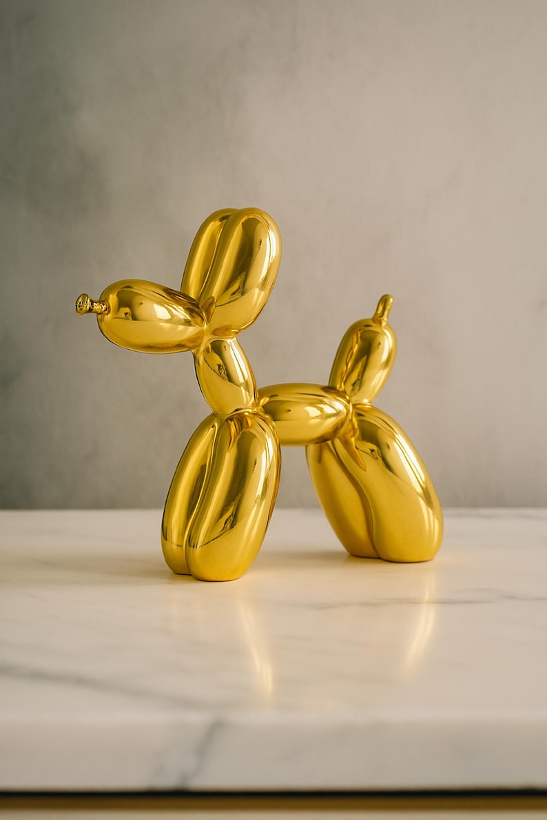 Op de afbeelding: Een gouden ballonhondsculptuur met een glanzende afwerking. De hond staat, zijn lichaam gevormd door verbonden ballonvormen. De sculptuur staat op een wit oppervlak tegen een neutrale achtergrond.