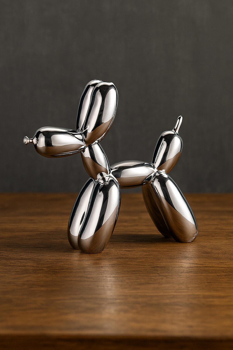 Op de afbeelding: Een gepolijste zilveren ballonhondsculptuur. De hond is gemaakt van glad, reflecterend materiaal, met een afgerond lichaam en langwerpige ledematen. De sculptuur wordt getoond op een houten oppervlak, met een donkere achtergrond.