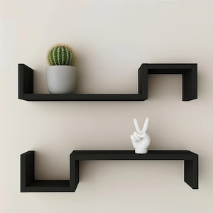 May include: Two black floating shelves with a modern design are mounted on a light beige wall. The top shelf holds a potted cactus, and the bottom shelf displays a white hand sculpture making a peace sign.