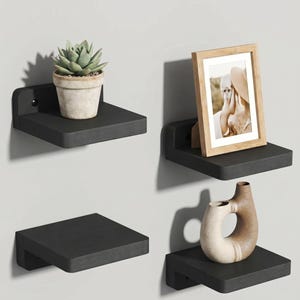 May include: Four black floating shelves mounted on a light gray wall. Each shelf displays a different decorative item: a potted succulent, a framed photograph, and a ceramic vase. The shelves are square with a curved bracket.