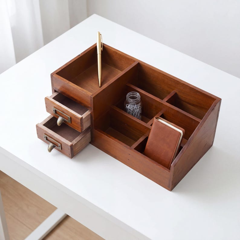 May include: A wooden desk organizer with multiple compartments and two small drawers. A gold pen rests in one section, and a small leather-bound notebook sits in another. The organizer is made of dark brown wood.