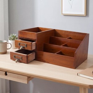 May include: A wooden desk organizer with multiple compartments and two small drawers. The organizer is made of dark brown wood and has metal label holders on the drawers. It sits on a light-colored wooden desk.