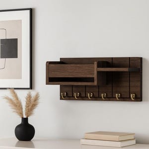 Op de afbeelding: Een donkerbruine houten sleutelhouder voor aan de muur, met een brievenbus en een plank. Het heeft zes messingkleurige haken voor sleutels. Het ontwerp is modern en minimalistisch, ideaal voor entree-organisatie.
