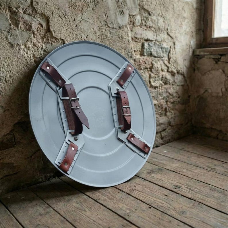 May include: A gray, round shield with brown leather straps and metal accents. The shield is leaning against a textured stone wall and wooden floor. The straps are secured with buckles. The shield has a classic, vintage design.