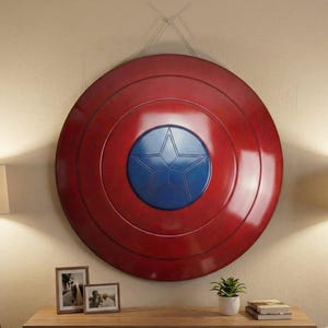 K&ouml;nnte beinhalten: Ein gro&szlig;er, runder Schild in Rot mit einer blauen Mittelscheibe und einem wei&szlig;en Stern. Der Schild h&auml;ngt an einer Wand und ist ein dekoratives Objekt. Es ist eine Nachbildung von Captain Americas Schild.