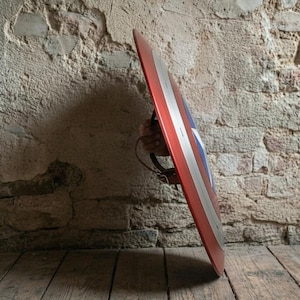 May include: A red, white, and blue shield with a star emblem and silver stripes. The shield is held by a hand, showcasing its design and color scheme. The shield is a prop or collectible.