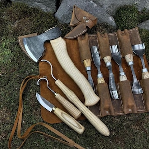 Puede incluir: Una colección de herramientas para trabajar la madera, que incluye un hacha, cuchillos y cinceles, dispuestas sobre un rollo de cuero marrón. Las herramientas tienen mangos de madera y hojas de metal. La escena está ambientada sobre musgo verde.