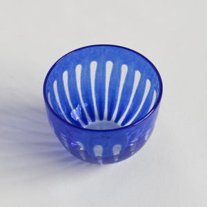 Puede incluir: Un pequeño cuenco redondo de cristal azul cobalto con diseños recortados blancos verticales. El cuenco es de cristal transparente y está sobre una superficie blanca. El diseño del cuenco recuerda a una flor o un estallido solar.