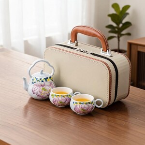 Op de afbeelding: Een witte keramische theepot en twee bijpassende theekopjes, elk versierd met een paars en groen bloemmotief. De set wordt getoond naast een beige, rechthoekige harde koffer met een bruin kunstlederen handvat en een ritssluiting.