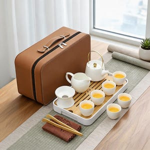 Puede incluir: Un juego de té de cerámica blanca que incluye una tetera con asa dorada, una jarra, un colador y seis tazas pequeñas sobre una bandeja rectangular de bambú, acompañado de un maletín de transporte marrón y pinzas de madera.