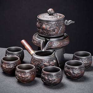 Könnte beinhalten: Dunkelgraues Keramik-Teeservice mit Drachenmotiv. Das Set beinhaltet eine Teekanne mit Deckel, eine Servierkanne und sechs kleine Teetassen. Das Drachendesign ist in einer kontrastierenden Bronzefarbe gehalten.