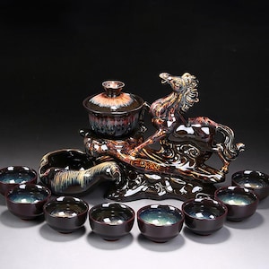 Puede incluir: Un juego de té de cerámica con una escultura de caballo. El juego incluye una tetera con tapa, un cuenco para servir y varias tazas de té pequeñas. Las piezas están esmaltadas en tonos marrones, negros y dorados, con interiores iridiscentes.