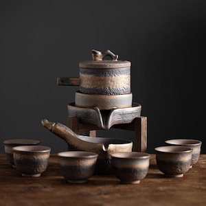 May include: A ceramic tea set with a rustic aesthetic. The set includes a teapot with a handle and lid, a serving tray, and several small teacups. The pieces are primarily brown and gold, with a textured finish. The set is displayed on a wooden surface.