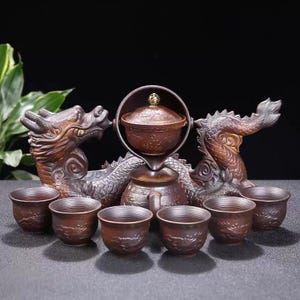 May include: A bronze-coloured tea set with a dragon design. The set includes a teapot, a serving vessel, and six small cups. Dragon sculptures flank the tea set, adding a decorative element. The tea set is made of metal.