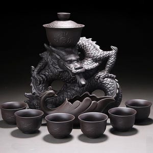 Op de afbeelding: Een donkergrijze keramische theeset met een drakenbeeld. De set bevat een theepot met deksel, een serveerschaal en zes bijpassende kopjes. De draak is ingewikkeld gedetailleerd en de kopjes hebben een subtiel ontwerp.