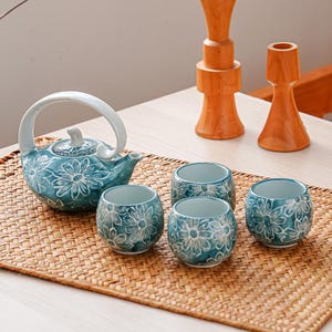 Puede incluir: Un juego de té de cerámica turquesa con un diseño floral, que incluye una tetera y tres tazas, se encuentra sobre una estera tejida. Dos candelabros de madera están en el fondo. La tetera tiene un asa y un pico blancos.