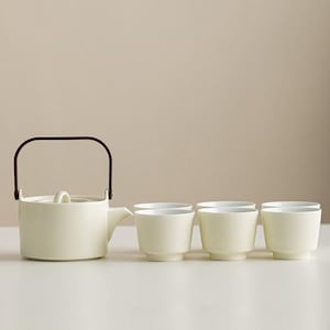 Puede incluir: Una tetera de cerámica color crema con asa y tapa marrón, acompañada de cuatro pequeñas tazas de té color crema. El conjunto está dispuesto sobre una superficie blanca sobre un fondo neutro, sugiriendo una estética minimalista.
