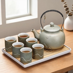 May include: A light green ceramic tea set, comprising a teapot and six cups, each filled with tea. The cups and teapot sit on a white and bamboo tray, placed on a wooden table. The teapot features a black handle and a mountain design.