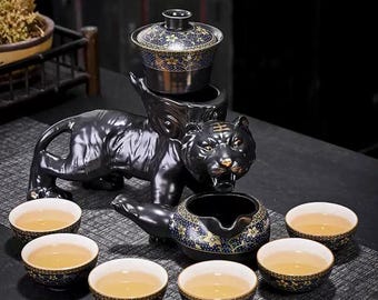 May include: A black ceramic tea set featuring a tiger-shaped base supporting a tea pot and a lidded serving bowl. Six small tea cups with floral patterns and tea are arranged around the set. The tea set has gold accents.