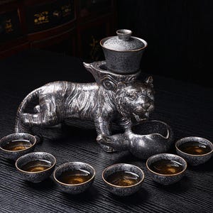 Peut inclure: Service à thé en céramique avec un support en forme de tigre argenté et noir. L'ensemble comprend une théière, une verseuse et six petites tasses à thé avec des bords dorés. L'ensemble est conçu pour servir le thé.