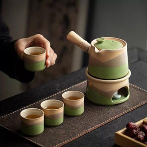 Puede incluir: Un juego de té de cerámica con una combinación de colores verde, beige y marrón. El juego incluye una tetera con asa de madera, un soporte para calentar y varias tazas pequeñas. Las tazas y la tetera tienen un diseño en capas. El juego de té está sobre una alfombra marrón.