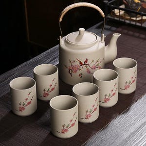 May include: A beige ceramic tea set featuring a teapot with a woven handle and six matching cups. The teapot and cups are decorated with pink floral and bird designs. The set is arranged on a dark brown surface.