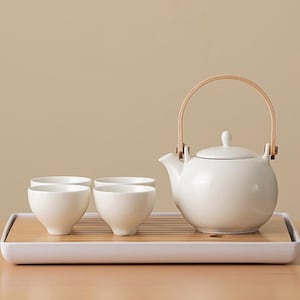 May include: A white ceramic tea set with a wooden handle on the teapot. The set includes four small, white teacups and a rectangular bamboo tray. The teapot and cups are arranged on the tray, ready for tea service.