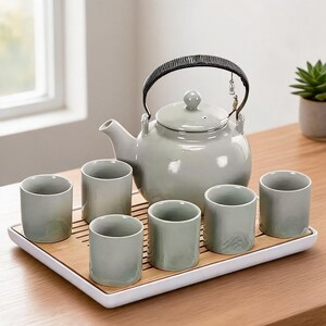 May include: A light grey ceramic teapot with a black handle and lid, accompanied by six matching cylindrical tea cups. The set is arranged on a rectangular white tray with a removable bamboo slatted insert for draining excess liquid.