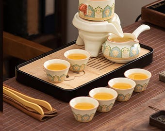 May include: A ceramic tea set with a decorative design. The set includes a tea pot, a serving pitcher, a tea warmer, and six small tea cups, all filled with tea. The set is on a bamboo tray.