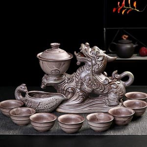 Könnte beinhalten: Ein Keramik-Teeservice mit einem drachenförmigen Mittelstück, einer Teeschale mit Deckel, einem schwanenförmigen Gefäß und mehreren kleinen Teetassen. Das Set ist hauptsächlich in einem gedämpften Braunton gehalten und weist aufwendige Details auf.
