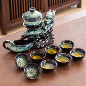 May include: A ceramic tea set with a dragon-shaped stand, a teapot, a lidded tea bowl, and several small teacups. The set features a teal and black glaze. The teacups contain a yellow liquid.