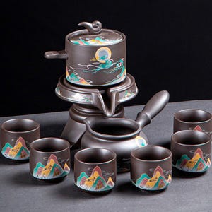 May include: A dark brown ceramic tea set. The set includes a teapot, server, and six cups. Each piece is decorated with a mountain landscape design in yellow, green, and red. The teapot has a lid and handle, and the cups are small.