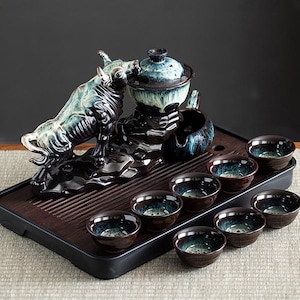 May include: A ceramic tea set featuring a bull figurine, a teapot with a lid, a small pitcher, and eight teacups. The set is primarily dark brown and teal, with a wooden tray. The bull is a prominent decorative element.