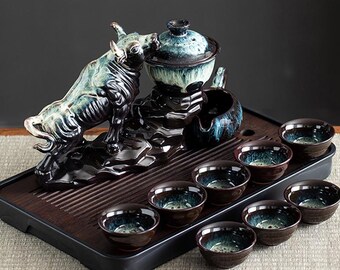 May include: A ceramic tea set featuring a bull figurine, a teapot with a lid, a small pitcher, and eight teacups. The set is primarily dark brown and teal, with a wooden tray. The bull is a prominent decorative element.