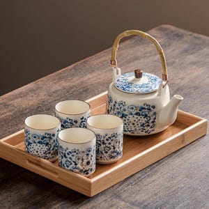 Pode incluir: Um conjunto de chá de cerâmica branca com desenhos florais azuis, incluindo uma chaleira com alça de bambu e quatro xícaras combinando. O conjunto é apresentado em uma bandeja retangular de bambu.