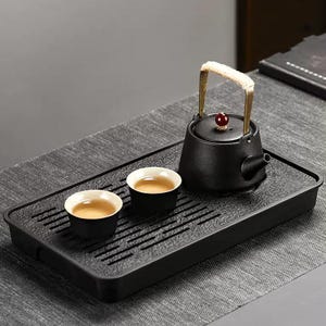 May include: A black tea set featuring a small teapot with a gold handle and a red knob, along with two small teacups. The set is placed on a rectangular black tray with a textured surface, perfect for serving tea.