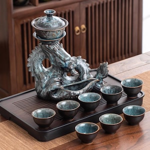 Puede incluir: Un juego de té en color verde azulado y bronce con un servidor en forma de dragón, una tetera con tapa y ocho tazas pequeñas. El juego se exhibe en una bandeja de madera oscura, con un mueble de madera en el fondo.