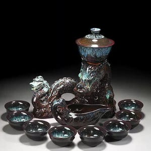 May include: A ceramic tea set with a dragon motif. The set includes a large teapot with a lid, a serving vessel, and several small teacups. The pieces are dark brown with blue and green accents.