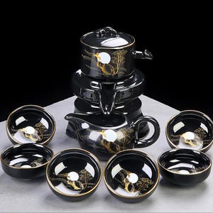 Op de afbeelding: Een zwart keramisch theeservies met gouden details. De set bevat een theepot, een serveerkan, een warmhoudstandaard en diverse kleine kommen. Elk stuk heeft een gouden boom- en maanontwerp. Het theeservies staat op een wit oppervlak.