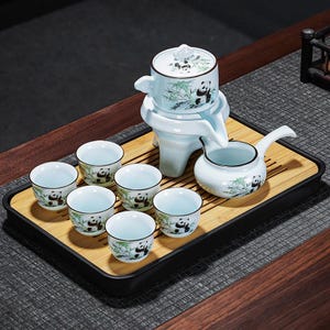 Könnte beinhalten: Ein hellblaues Keramik-Teeservice mit Panda-Bär- und Bambusmotiven. Das Set umfasst eine Teekanne, eine Servierkanne und sechs kleine Tassen, alles auf einem Bambustablett angeordnet. Das Teeservice ist zum Servieren von Tee bestimmt.