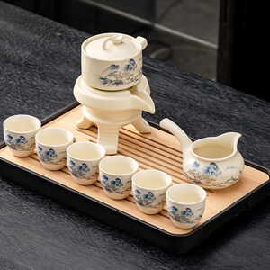 May include: A cream-coloured ceramic tea set, including a teapot with lid, serving pitcher, and six small teacups. Each piece is decorated with a blue and white landscape design. The set is presented on a bamboo tray.