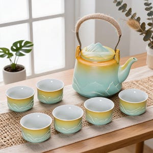 Pode incluir: Um conjunto de chá em cerâmica composto por um bule com pega revestida a corda e seis chávenas a condizer. As peças apresentam um acabamento em gradiente, do amarelo ao verde claro, com desenhos em relevo de cadeias montanhosas.